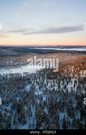 Aerial view of a vast snow-covered coniferous forest in the Arctic landscape, illuminated by the warm light of the setting sun with a distant lake in Stock Photo