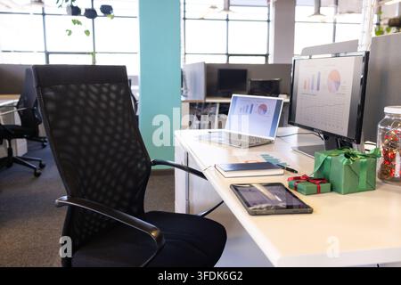 Ergonomic black chair occupying left side, by white desk displaying laptop, charts, gift boxes Stock Photo