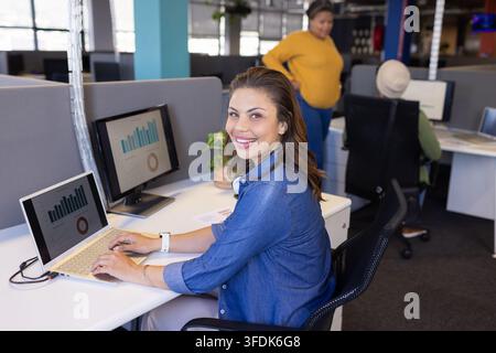 Woman smiling and typing on laptop at modern open plan office with monitors showing charts Stock Photo