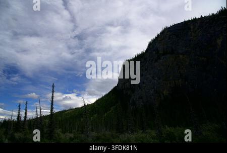 Dramatic view of a massive dark rock cliff and steep mountain slope lined with pine trees under a dynamic blue sky with thick white clouds Stock Photo