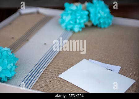 A close-up shot of a white paper envelope pinned to a rustic brown burlap backing board, surrounded by decorative turquoise blue paper flower clusters Stock Photo