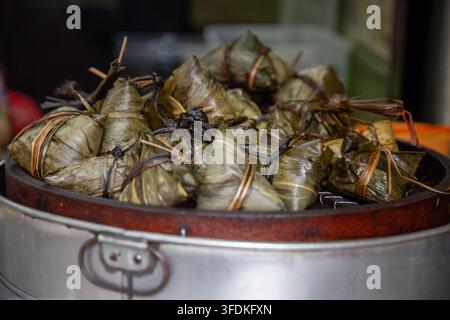 A mound of traditional zongzi—sticky rice dumplings wrapped carefully in dark green bamboo or reed leaves—resting inside a large metal steamer tray at Stock Photo