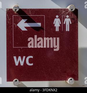 Burgundy toilet sign featuring a white arrow pointing left, alongside male and female pictograms and the WC abbreviation, providing clear directional Stock Photo