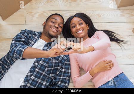 Loving black couple making heart with their hands, lying on floor surrounded by boxes on relocation day, top view Stock Photo