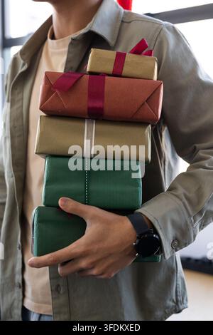 Asian man holding stack of 5 wrapped Christmas gifts by window in khaki shirt showing smartwatch Stock Photo