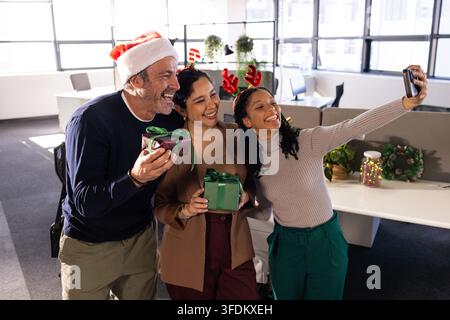 Diverse coworkers taking selfie at office using phone, wearing Santa hats, holding Christmas gifts Stock Photo