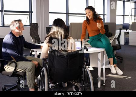 Diverse coworkers collaborating around round table in bright office with laptop and wheelchair Stock Photo