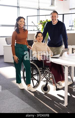 Diverse coworkers collaborating around white table in open office with manual wheelchair and tablet Stock Photo
