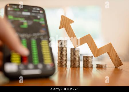Mobile stock trading and FinTech investment concept with smartphone displaying market chart beside coin stacks and wooden upward trend arrow. Stock Photo