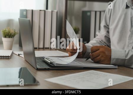 Businessman checking payroll documents on clipboard beside laptop for payroll processing, salary review, employee records, HR administration, accounti Stock Photo