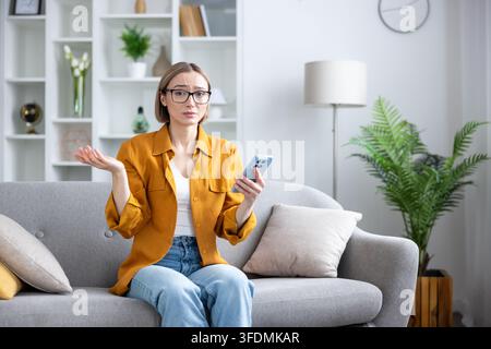 Woman sitting on a sofa in a modern living room, holding a smartphone, looking at the camera with a puzzled expression and shrugging, representing confusion, doubt, or digital frustration Stock Photo