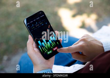 Woman is sitting on a bench, holding a smartphone displaying stock market data, rising chart and pressing the sell button. Woman is sitting on a bench Stock Photo