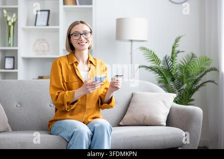 Woman sitting on sofa and smiling at camera while holding a smartphone and credit card, enjoying convenient internet shopping and secure digital payment Stock Photo