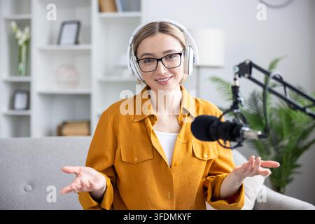 Woman wearing headphones and glasses smiling at camera while recording a podcast or streaming video content, gesturing with hands in her home studio setup Stock Photo
