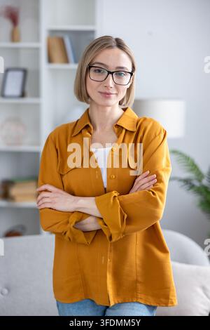 Young blonde woman with short hair and black framed glasses wearing a yellow casual shirt and blue jeans, standing with arms crossed in a modern home office or living room Stock Photo