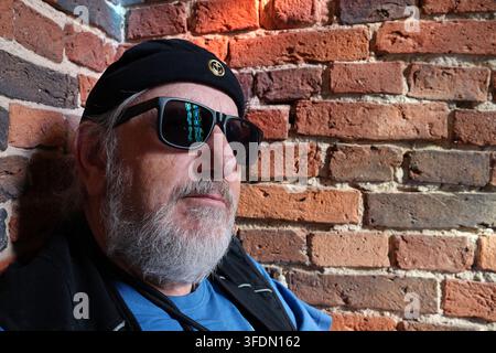Elderly man with a beard wearing dark sunglasses and a black beanie with a small anchor emblem. He is dressed in a blue shirt and a black vest. The ba Stock Photo