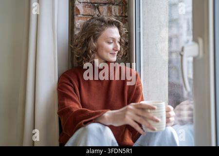 Calm satisfied woman freelancer taking pause from remote work for stress free at home. Relaxed dreamy young female enjoying mindful break on windowsil Stock Photo