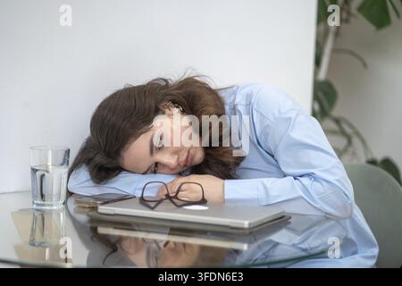 Exhausted young woman resting head on table near laptop, eyeglasses. Concept of burnout, mental fatigue, emotional exhaustion, work stress, lack of mo Stock Photo