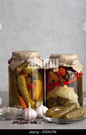 Two large jars of fermented pickles and mixed vegetables, surrounded by garlic bulbs, a small glass bowl of seasoning, and pickled cucumbers on a plat Stock Photo