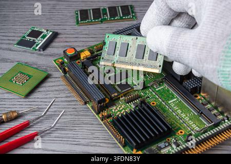 A technician is seen working on a computer motherboard in a workshop. They are carefully handling memory chips and using tools. Stock Photo