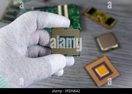 A person in a glove is shown holding a computer chip. Several other chips and circuit boards are visible on a gray work table. Stock Photo