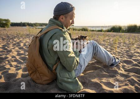 Cat travel. Relaxed traveler with backpack enjoys rest with cat on beach. Thoughtful man sitting with kitty on laps outdoor emotionally connected with Stock Photo