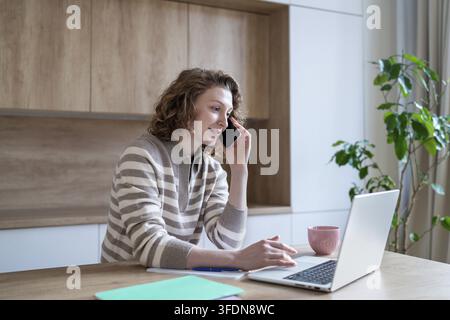 Focused interested woman remote working from home, skilled multitasking between business phone call and laptop. Confident young female freelancer mana Stock Photo