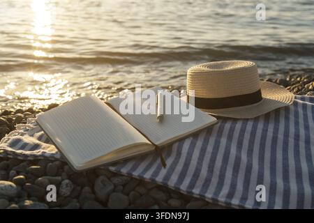 Open notebook with pen resting on striped beach blanket near calm sea water at golden sunset, straw hat beside journal creating peaceful summer vacati Stock Photo