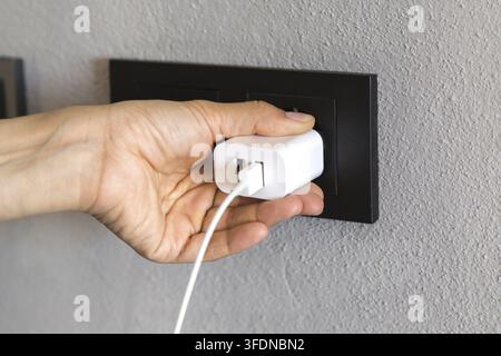 Human hand plugging white USB charger with cable into black electrical socket on gray wall, symbolizing energy use, technology connection, charging pr Stock Photo