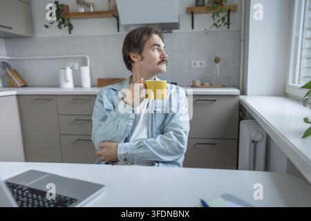 Calm reflective man holding mug of coffee in home kitchen, looking away deep in thought. Pensive young man with cup in hand taking break at table, gaz Stock Photo