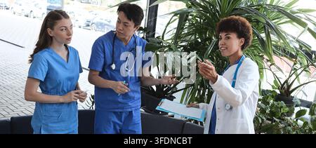 Multiracial medical team engaged in an active clinical debriefing session surrounded by green plants in a bright hospital lounge Stock Photo