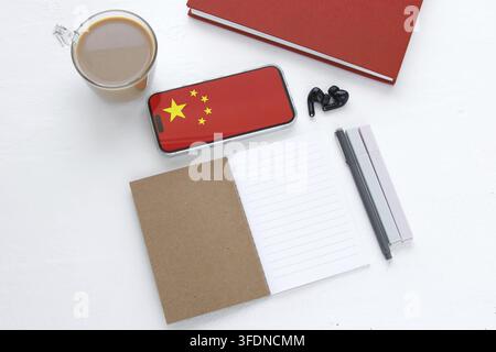 Flat lay with smartphone showing Chinese flag, notebook, pens, coffee cup and earbuds on white desk, symbolizing learning Chinese language, education Stock Photo