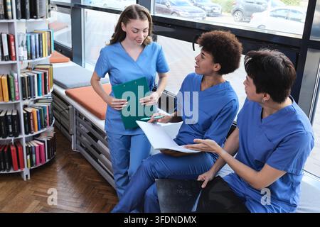 Elevated view of three multicultural interns in uniform collaborating on complex clinical data surrounded by library book racks Stock Photo