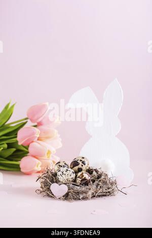 Happy easter quail eggs in a nest, white rabbit and tulips on a pink background vertical view Stock Photo