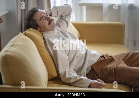 A tired, exhausted man who looks into the void feels deep tiredness, life pressure and burnout while sitting on the sofa Stock Photo