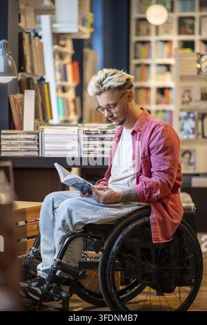 Focused young man with disability reading book in bookstore. Interested student guy in wheelchair wearing eyeglasses holds textbook in modern library. Stock Photo