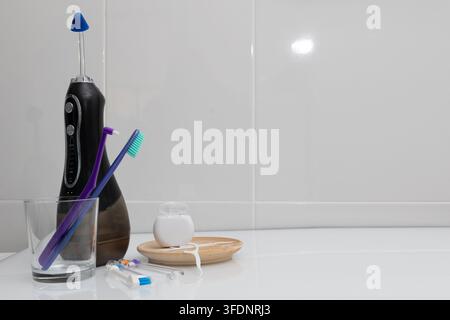 A left dominant cluster of home dental care tools including electric and manual toothbrushes, a glass cup, and a wooden coaster on a bathroom counter Stock Photo
