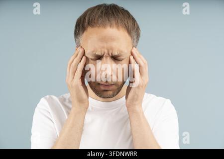 Unhealthy frowning man suffers from intense migraines, severe headaches, holding head with pulsating pain. Symptoms pounding in temples by stress, deh Stock Photo