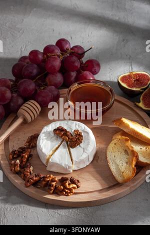 Cheese plate, still life of seasonal wine snacks. Camembert or brie cheese, grapes, walnuts, baguette, honey and figs with interesting shadows in the Stock Photo