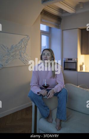 Interested serene woman rest on sofa with glass of red wine, watching movie in cozy apartment. Pleased smiling mature female enjoy relaxed evening wit Stock Photo