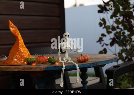 A small plastic Halloween skeleton figurine sits on a wooden veranda table, raises one arm as if waving, surrounded by pumpkins, a witch hat, and stri Stock Photo