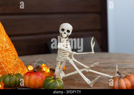 Playful plastic Halloween skeleton figurine sits on a wooden tabletop with warm left lighting, orange string lights, pumpkins, and cobweb fabric, crea Stock Photo