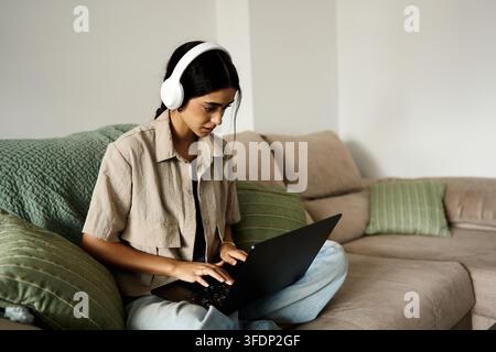Young woman focused on her work or e learning, comfortably sitting on a couch at home using a laptop and headphones Stock Photo