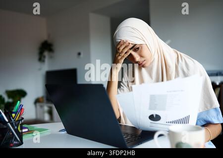Young muslim woman wearing a hijab feeling stressed while working on her laptop and reviewing financial documents in her home office Stock Photo