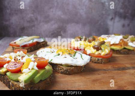 Freshly made whole grain toasts topped with avocado, eggs, tomatoes, salmon, mushrooms, and herbs on a wooden board Stock Photo