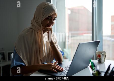 Muslim woman wearing a hijab smiling while typing on a laptop, working or studying from a home office near a window Stock Photo