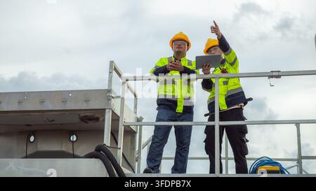 Two Asian male electrical engineers wearing hardhats and safety vests use tablet and walkie-talkie while pointing ahead on a high platform structure. Stock Photo
