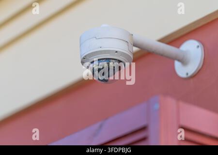 A sleek surveillance camera is mounted on a colorful wall observing the lively street below. Sunlight gleams off its shiny lens reflecting urban life Stock Photo