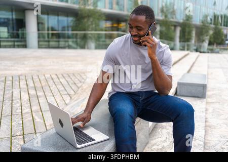 African American man freelancer using laptop talking on phone on urban street in city. Business man having virtual meeting. Professional programmer using phone laptop working outdoors. Remote work Stock Photo