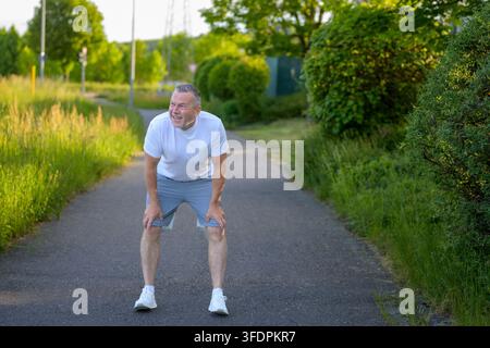 Senior man taking a short recovery break after jogging outdoors on a sunny pathway. Authentic healthy aging, endurance and active lifestyle concept wi Stock Photo